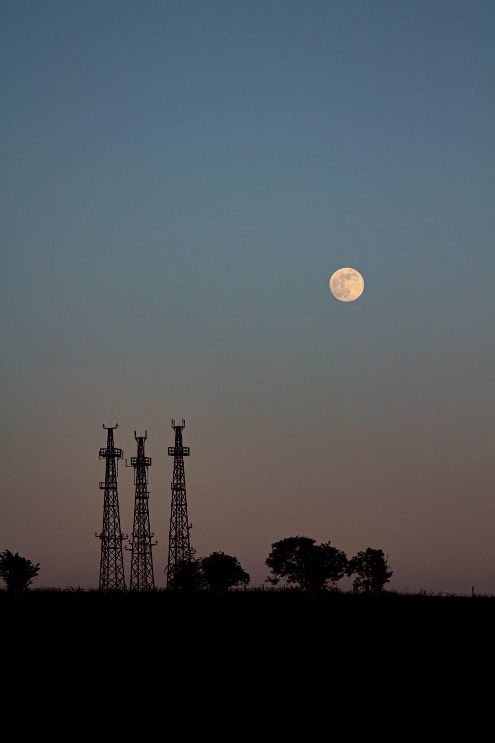 Landscape shot at dusk where the sky is an orange to blue gradient with the moon visible. On the horizon are some small trees and electrical towers