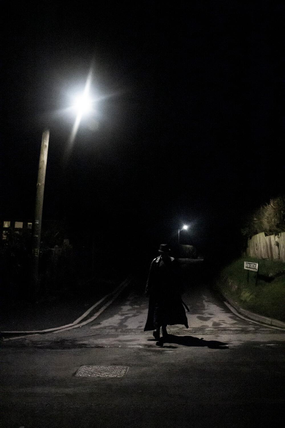 A shadowy figure walking toward the camera at night with a street light behind, casting harsh shadows